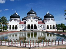 The white Baiturrahman Grand Mosque with black domes reflected in a foreground pool.