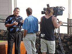 A man in a blue shirt, in front of a woman manning a camera, both engaging with a man in a black and blue shirt.