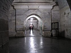 Vista interior de um túnel de pedra com vários arcos e alvenaria branca ornamentada, com visitantes caminhando por um corredor, um piso escuro brilhante refletindo as luzes do teto e um quadro de informações à direita.