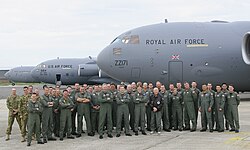 A group of people wearing overalls standing together in front of three grey aircraft are visible in the background.