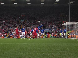 People in blue and red shirts on a field with a ball in the air. In the background is a stand that contains a lot of people.
