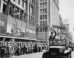 Patton en uniforme se tient debout dans un véhicule militaire. Des spectateurs saluent son passage tandis que des cotillons sont jetés depuis les gratte-ciels bordant la rue.