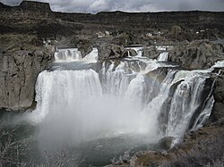 A wide, multi-stage waterfall, backed by dark cliffs, cascades into a rocky canyon