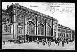 Carte postale montrant partiellement et en perspective la gare de Strasbourg, un imposant bâtiment de style wilhelmien, avec son entrée par la partie centrale et son aile droite. Ses façades, librement inspirées du style Renaissance, s'ouvrent par de grandes baies vitrées. Devant l'auvent de la gare stationnent plusieurs personnes et chevaux attelés ou montés. Les impressions en allemand Strassburg et Zentralbahnhoff figurent au-dessus de la gare parmi d'autres mots manuscrits.