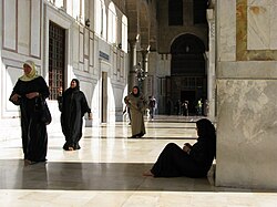 In a mosque several women wearing hijabs are walking, with one woman wearing hijab leaning against a pillar, a couple more people are in the background.