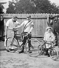 Photographie en noir et blanc d'un homme portant son vélo et d'un petit garçon avec une petite bicyclette, entourés de deux autres hommes.
