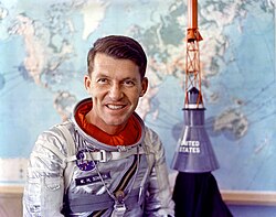 Schirra, sans casque mais en combinaison argentée, devant une maquette d'un engin spatial.