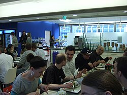 Cette photo en gros plan de l’angle d’une cantine en libre-service montre des gens attablés, une partie de comptoir, des armoires frigos pour les boissons. Le bleu roi des murs domine.