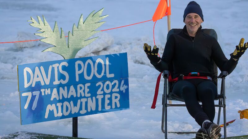 A man wearing all black, including a beanie, sits in a chair smiling, with a sign saying: Davis Pool 77th ANARE 2024 Midwinter