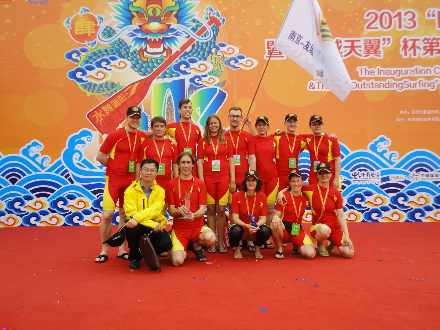 Nanjing 2013 Drachenboot Nanjing und Suzhou Mannschaftsfoto der Leipziger Drachenbootfahrer in roten Mannschaftsoutfit auf dem roten Teppich, im Hintergrund ein orange-blaues Werbeplakat für das Drachenbootfestival