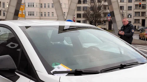 Weißer Pkw mit Parkverstoß-Zettel unter dem Scheibenwischer; im Hintergrund eine Parkkontrolleur.