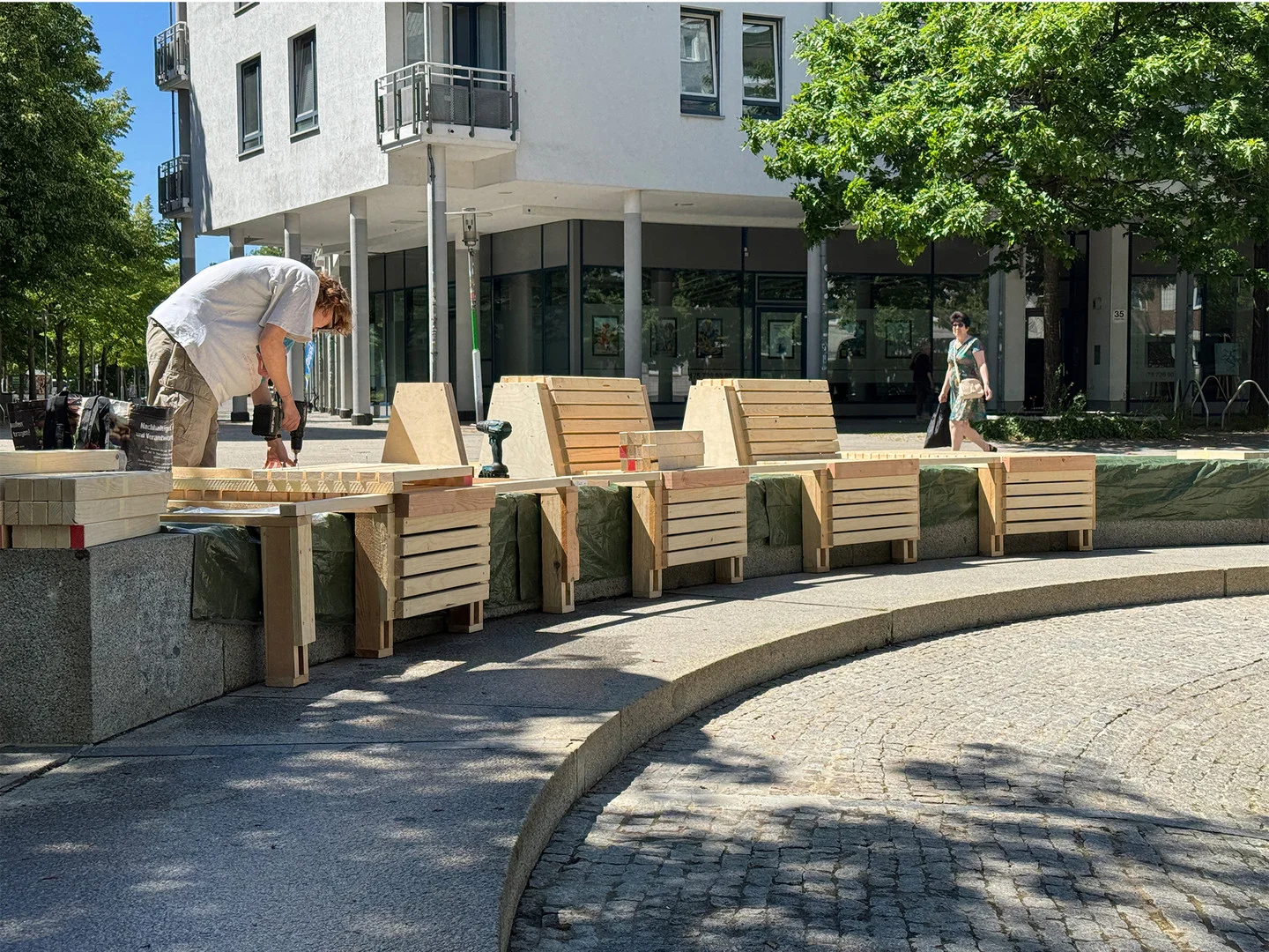 • Eine Frau montiert Holzlatten an der neuen Brunnenbank, im Hintergrund läuft eine Frau über den Platz.