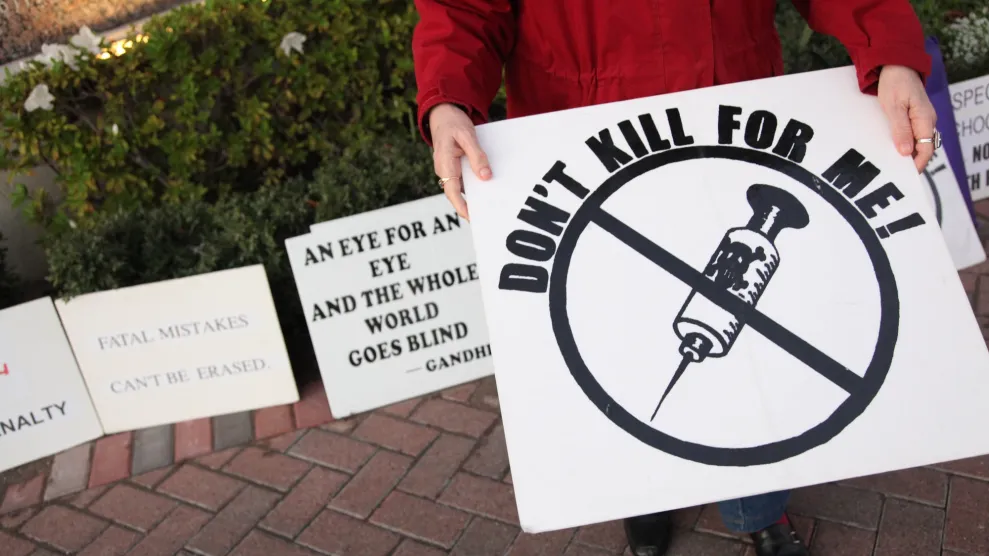 A person in a red jacket holding a sign that says "Don't Kill For Me" with a picture of a needle with a skull and crossbones crossed out