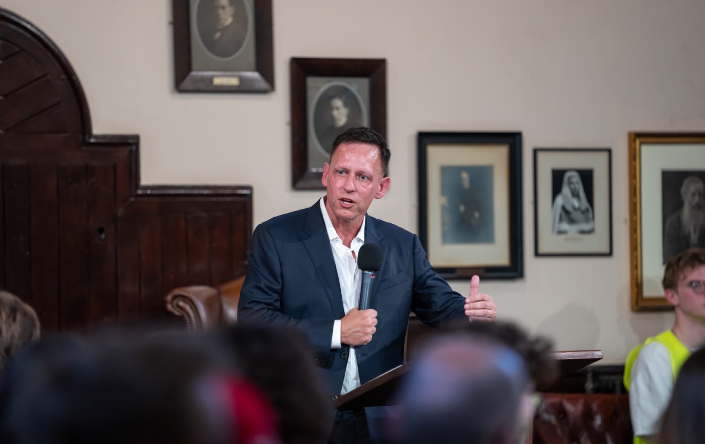 Peter Thiel speaks at a lectern, holding a microphone in front of the audience at The Cambridge Union.