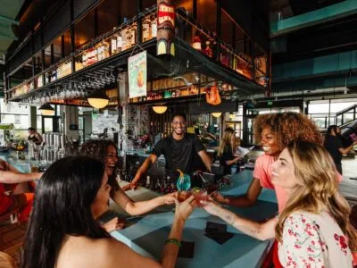 The bar scene at Moxy, featuring four friends toasting with colorful cocktails, while a smiling bartender engages with them in a vibrant atmosphere.