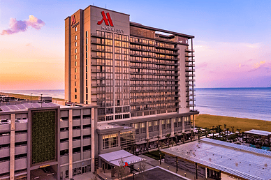 Aerial view of a modern Marriott hotel with ocean views and sunset colors in the background, featuring a parking garage nearby.