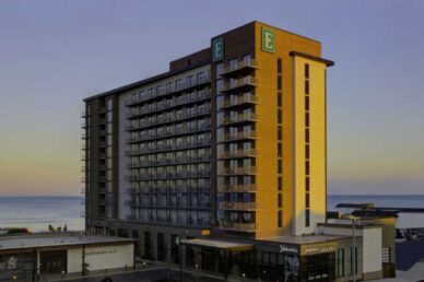 Modern beachfront Embassy Suites hotel with multiple floors, featuring balconies and a well-lit facade during sunset.